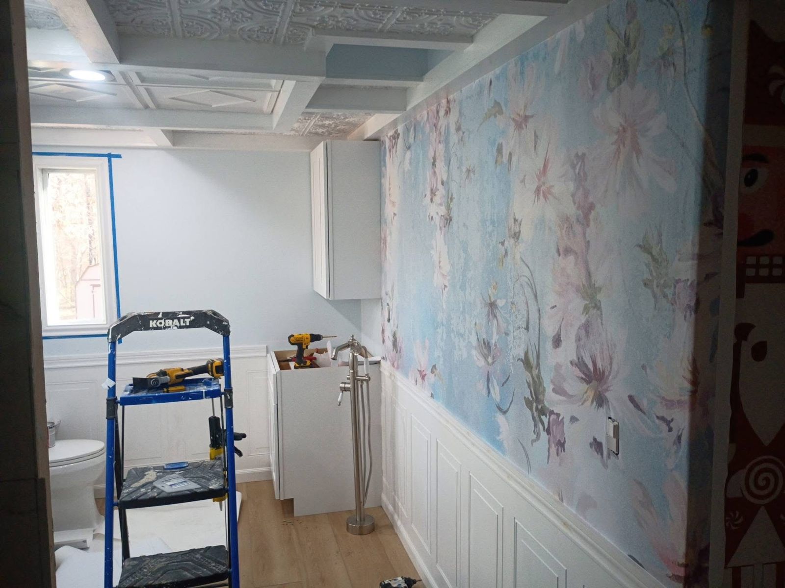 The remodeled bathroom coming together: floral mural above white wainscoting, coffered ceiling, new cabinets, wood-look floor and a freestanding tub filler